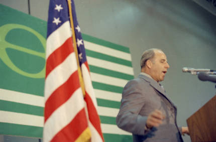 Gaylord Nelson speaking at a podium during Earth Week with an American flag in the foreground, and the Earth Day flag behind it.